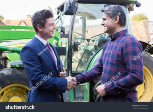 stock-photo-farmer-and-businessman-shaking-hands-with-tractor-in-background-489088585